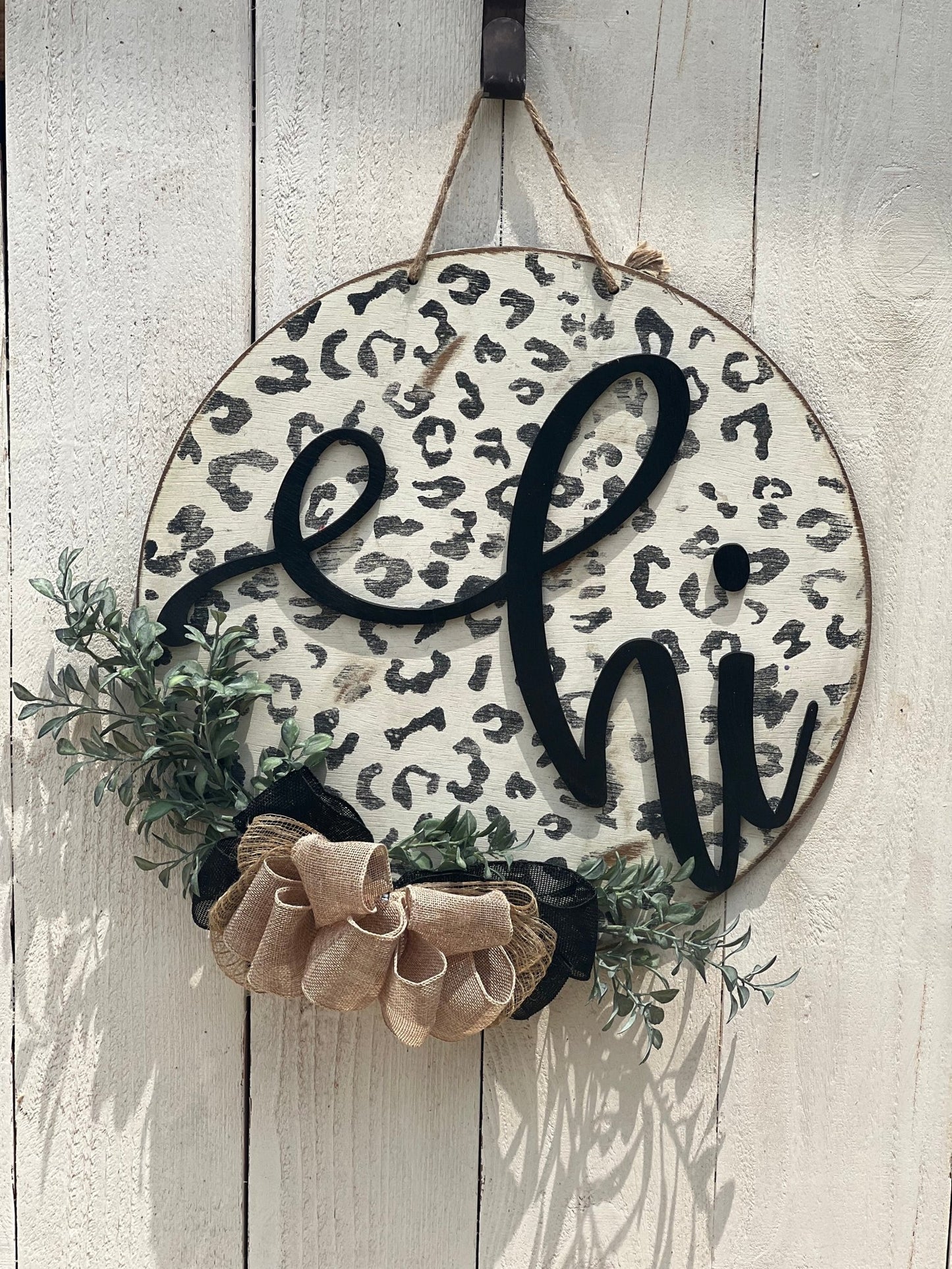 White leopard print door hanger made of wood, featuring a round shape and rustic-modern style on white fence.