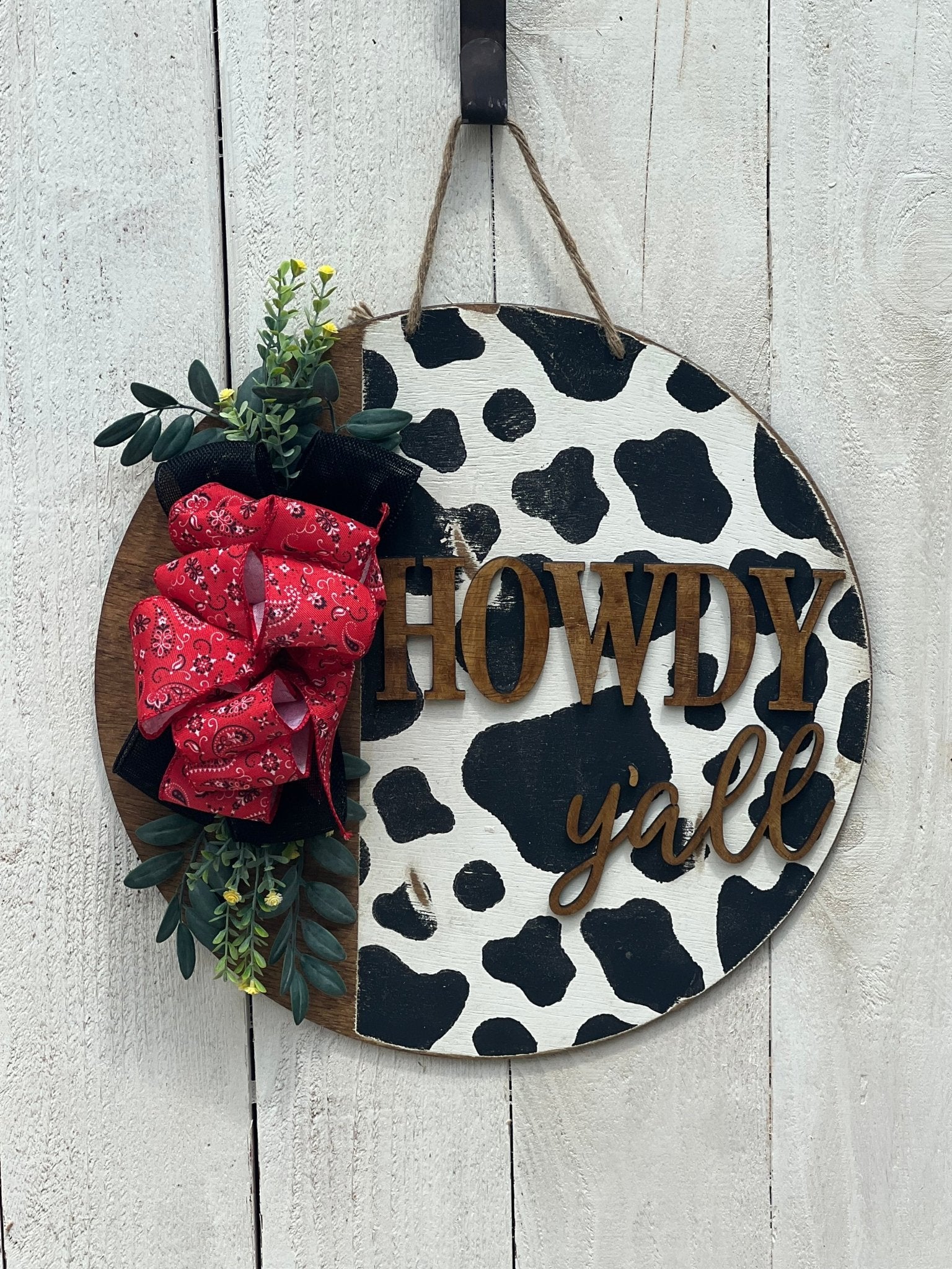 Cow Print Howdy Y’all Wood Door Hanger in black, red, and white with rustic charm hanging on a white fence.