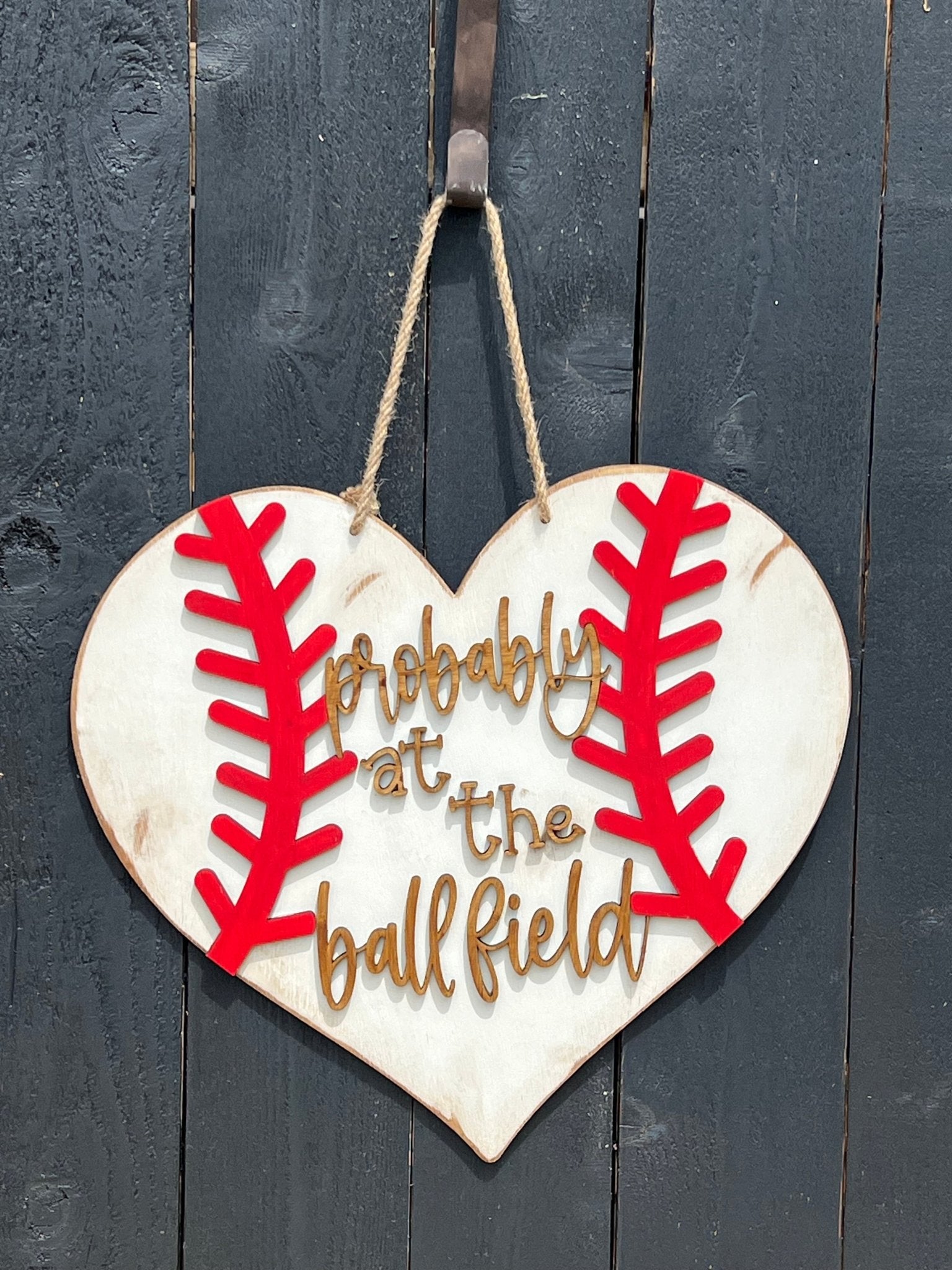 A rustic wooden door hanger shaped like a heart with baseball stitching in white and red on black fence.