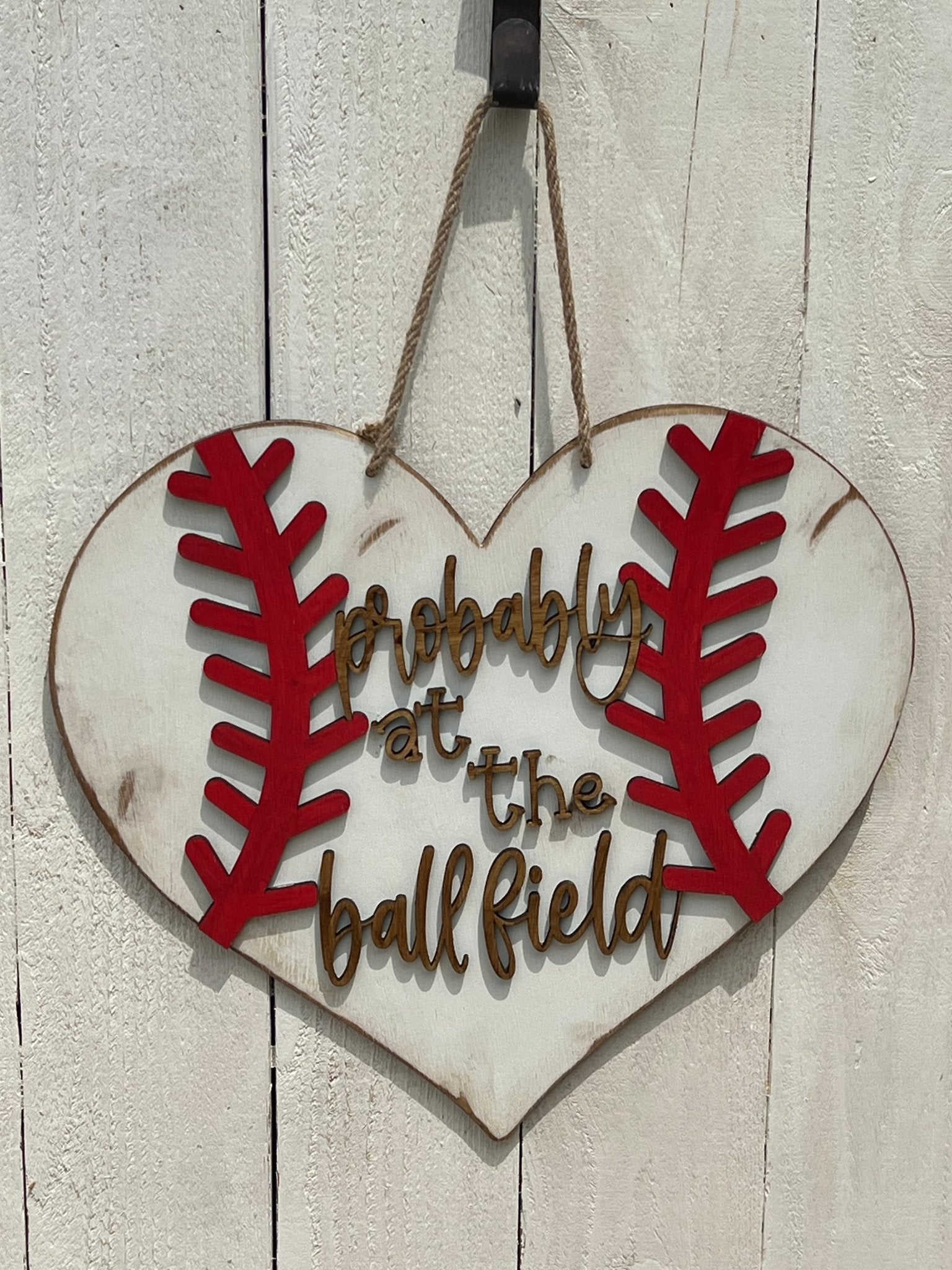 A rustic wooden door hanger shaped like a heart with baseball stitching in white and red on white fence.