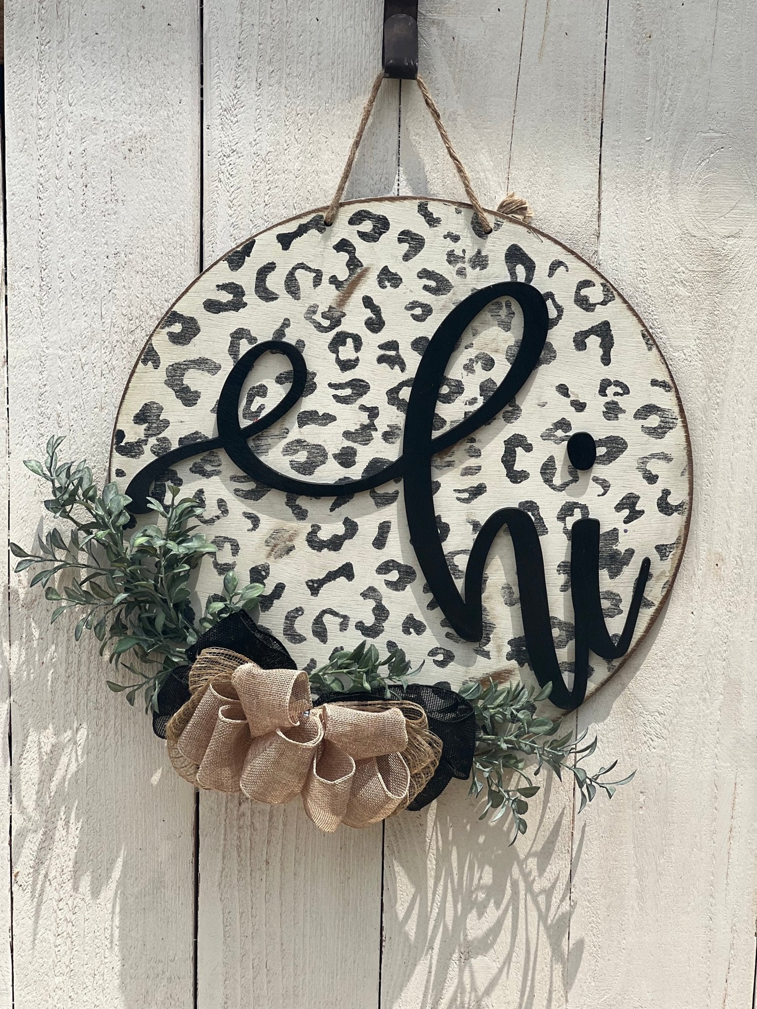 White leopard print door hanger made of wood, featuring a round shape and rustic-modern style on white fence.