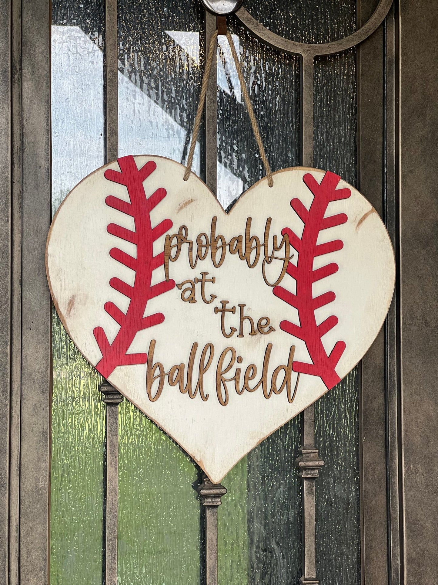 A rustic wooden door hanger shaped like a heart with baseball stitching in white and red on metal door.
