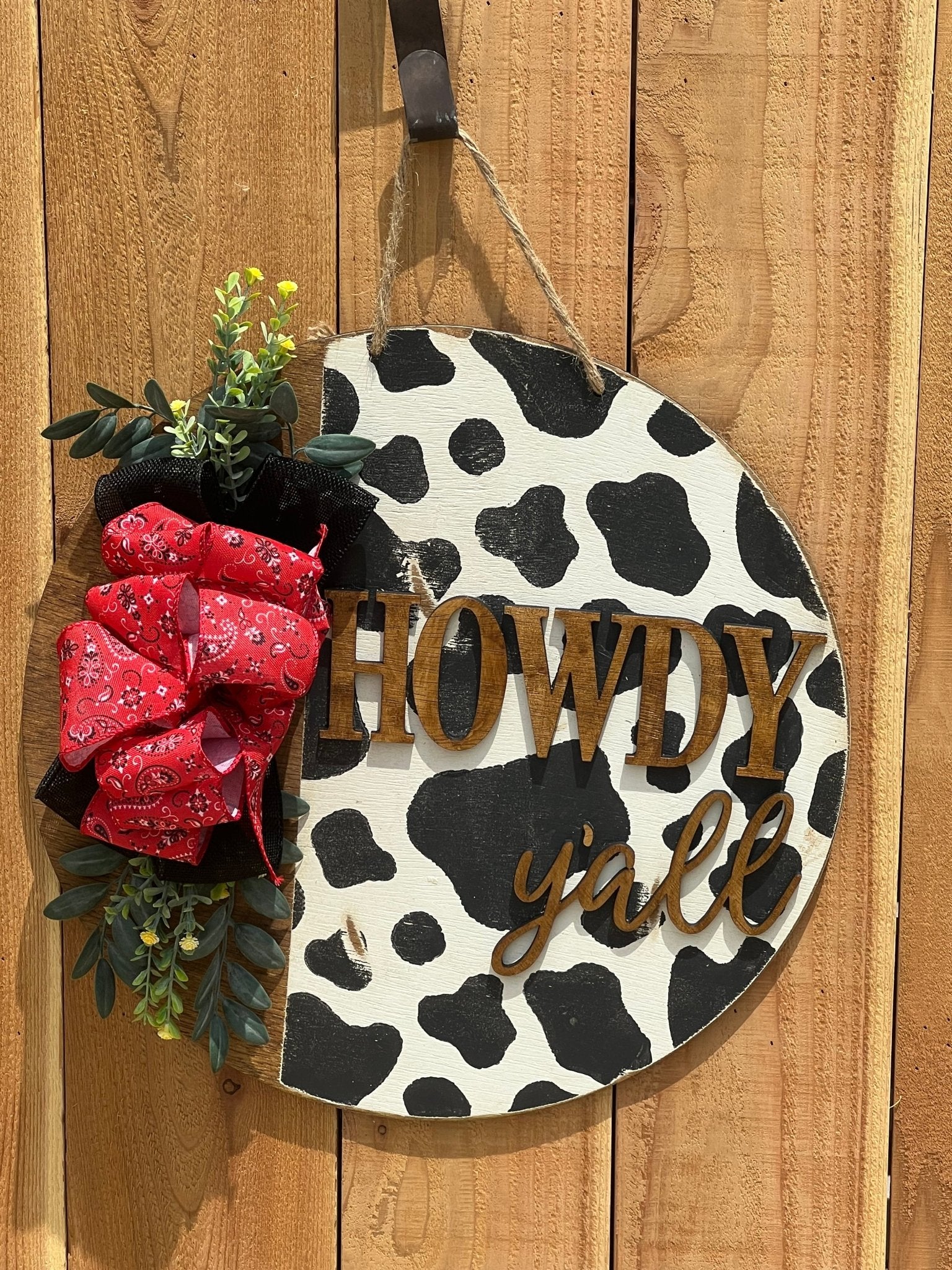 Cow Print Howdy Y’all Wood Door Hanger in black, red, and white with rustic charm hanging on a unpainted fence.