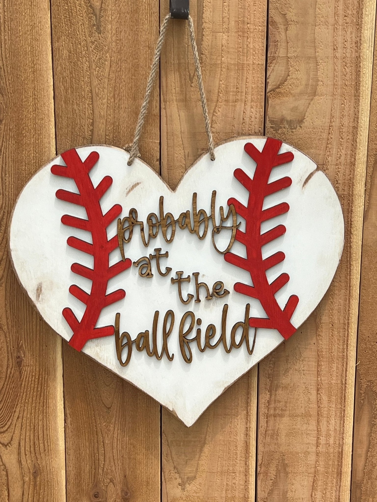 A rustic wooden door hanger shaped like a heart with baseball stitching in white and red on unpainted fence.