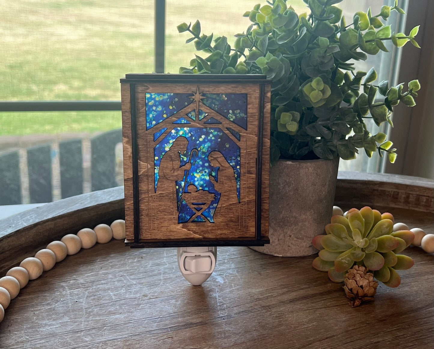 A Christmas Nativity Lantern Nightlight in golden oak with wood and acrylic materials on a try with natural light shining through.