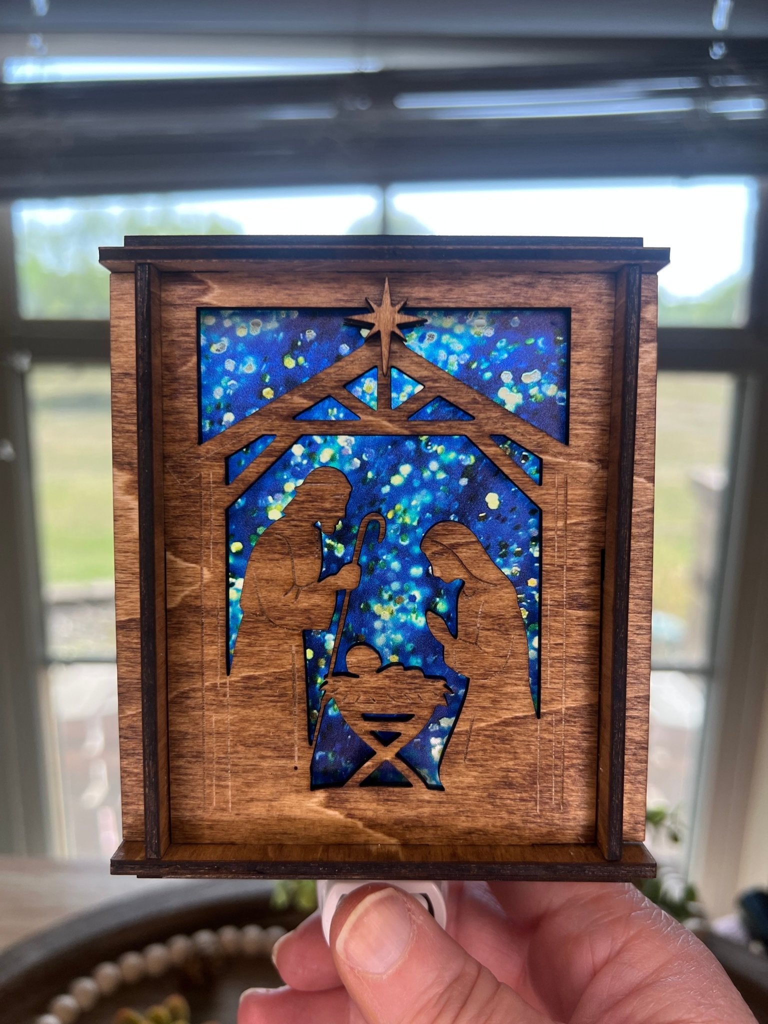 A Christmas Nativity Lantern Nightlight in golden oak with wood and acrylic materials held in hand.