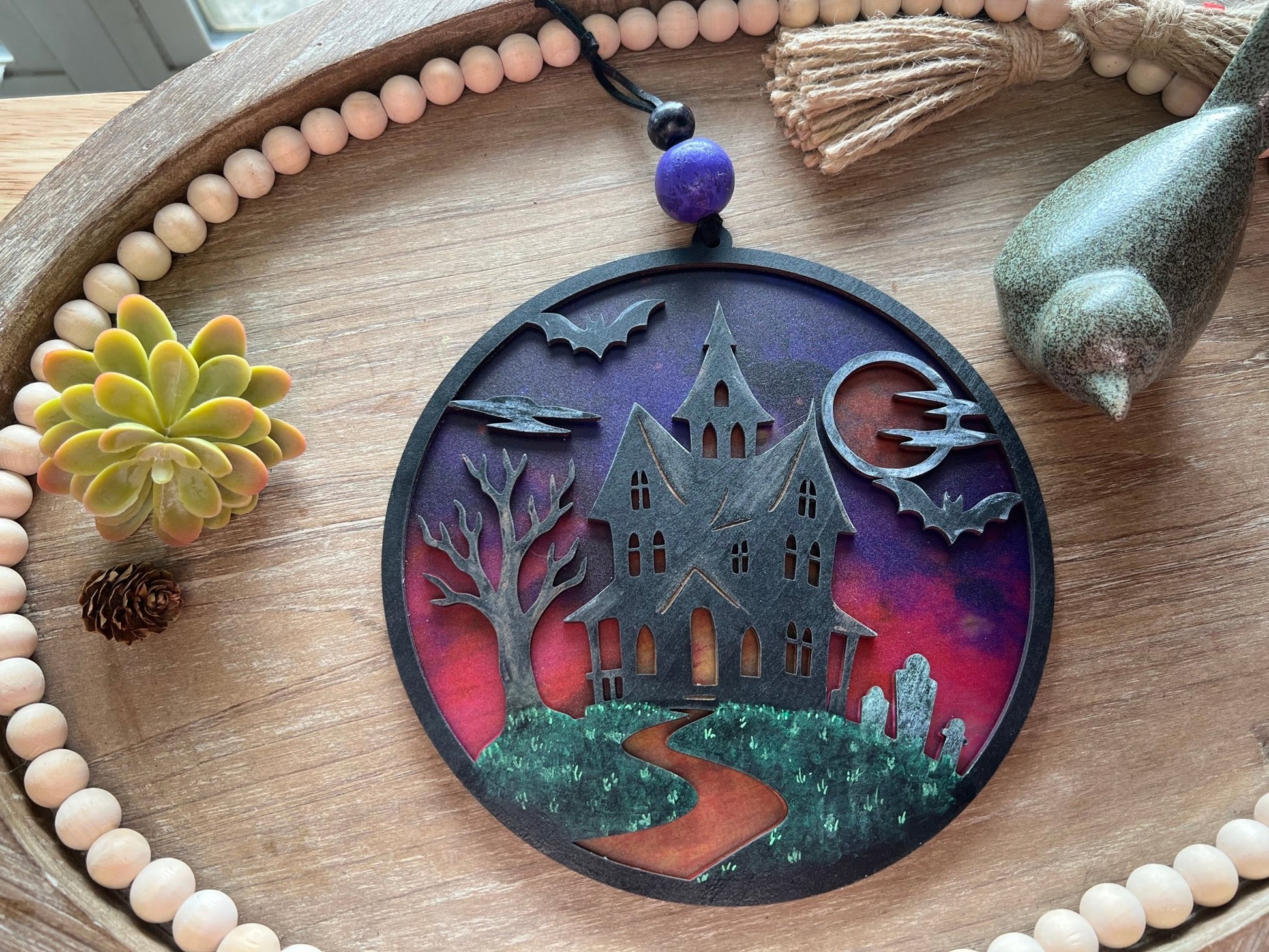 Hand painted suncatcher with a haunted house design on wood and acrylic, in vibrant colors laying on a try.