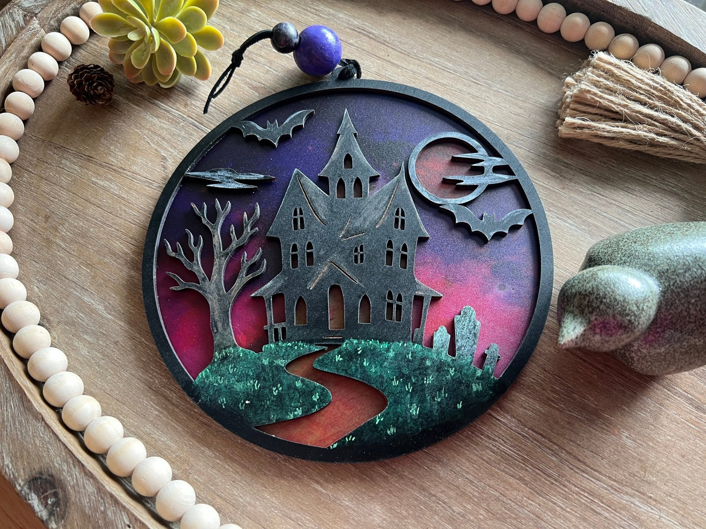 Hand painted suncatcher with a haunted house design on wood and acrylic, in vibrant colors - lying on a tray.