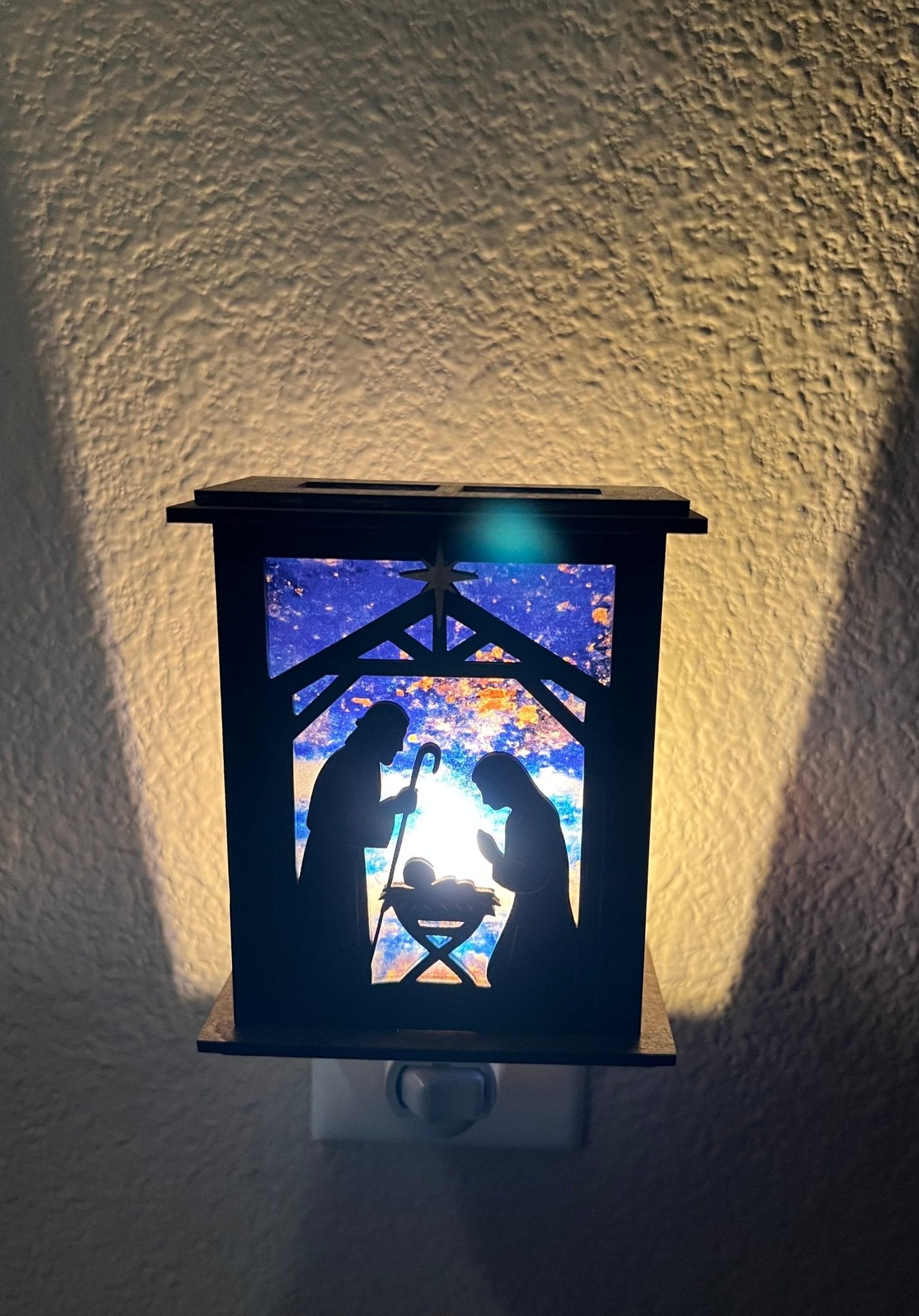 Rustic nativity nightlight lantern made of black wood and golden oak stained acrylic plugged in with front view and lit.