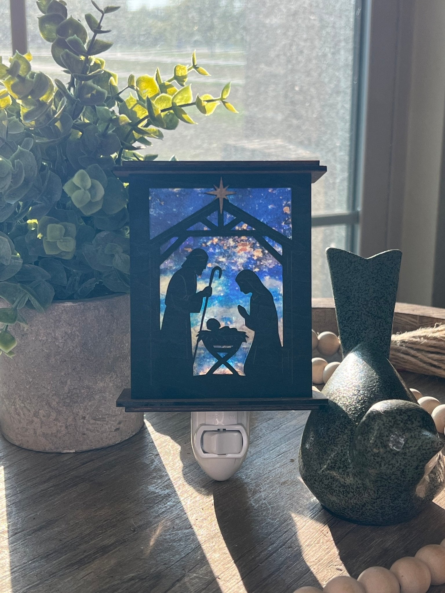 Rustic nativity nightlight lantern made of black wood and golden oak stained acrylic standingup  with front view and sunlit.